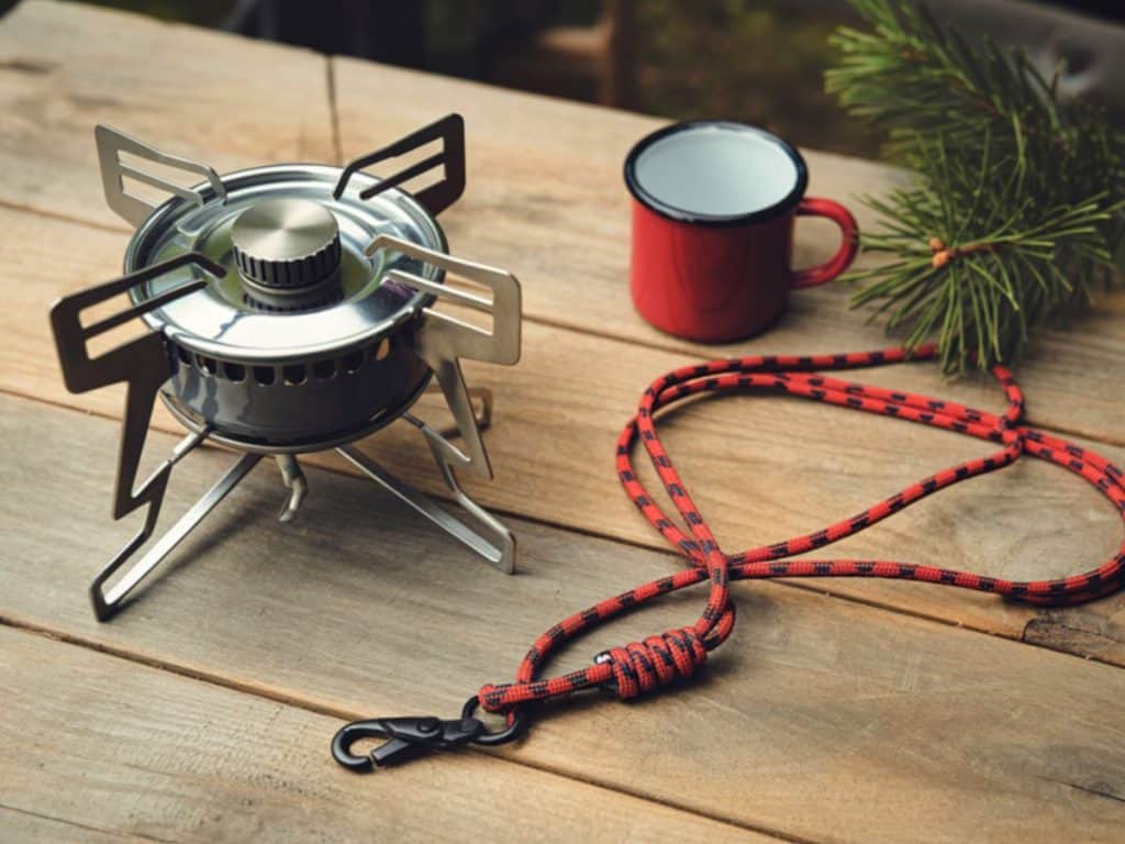 Lightweight backpacking gas stove on a wooden table beside a camping mug and paracord, highlighting a compact option for the best camping stove for hiking and backpacking.