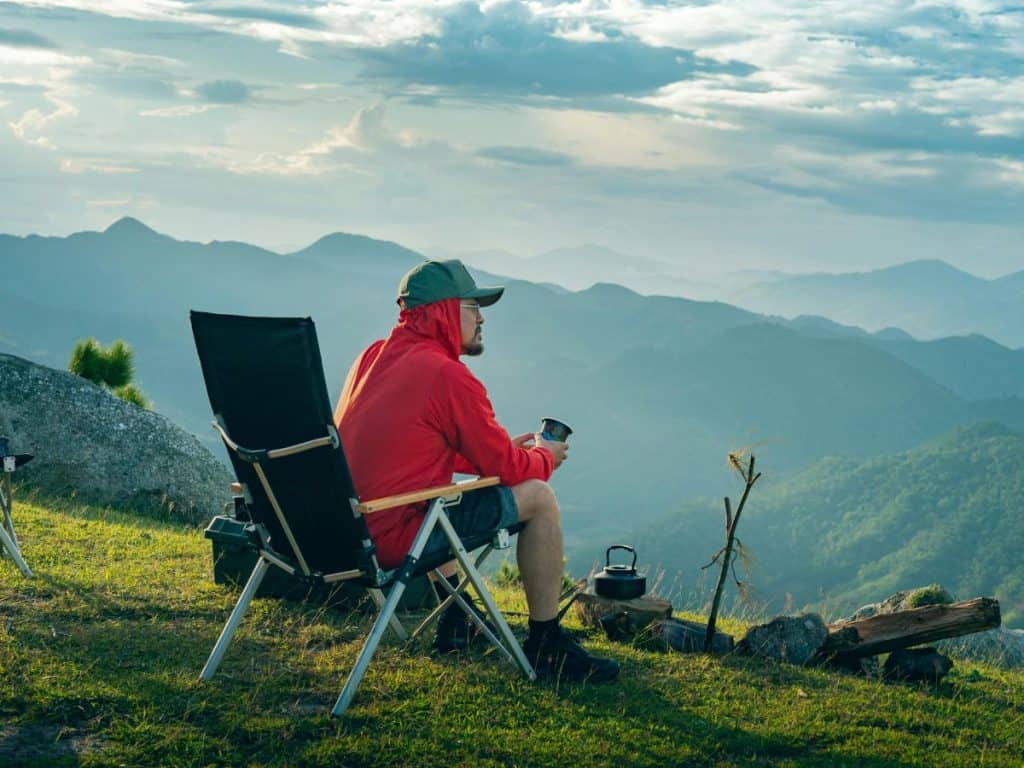 Camper relaxing on a mountainside with a hot drink beside a compact outdoor stove, showing the best camping stove for scenic campsite cooking.