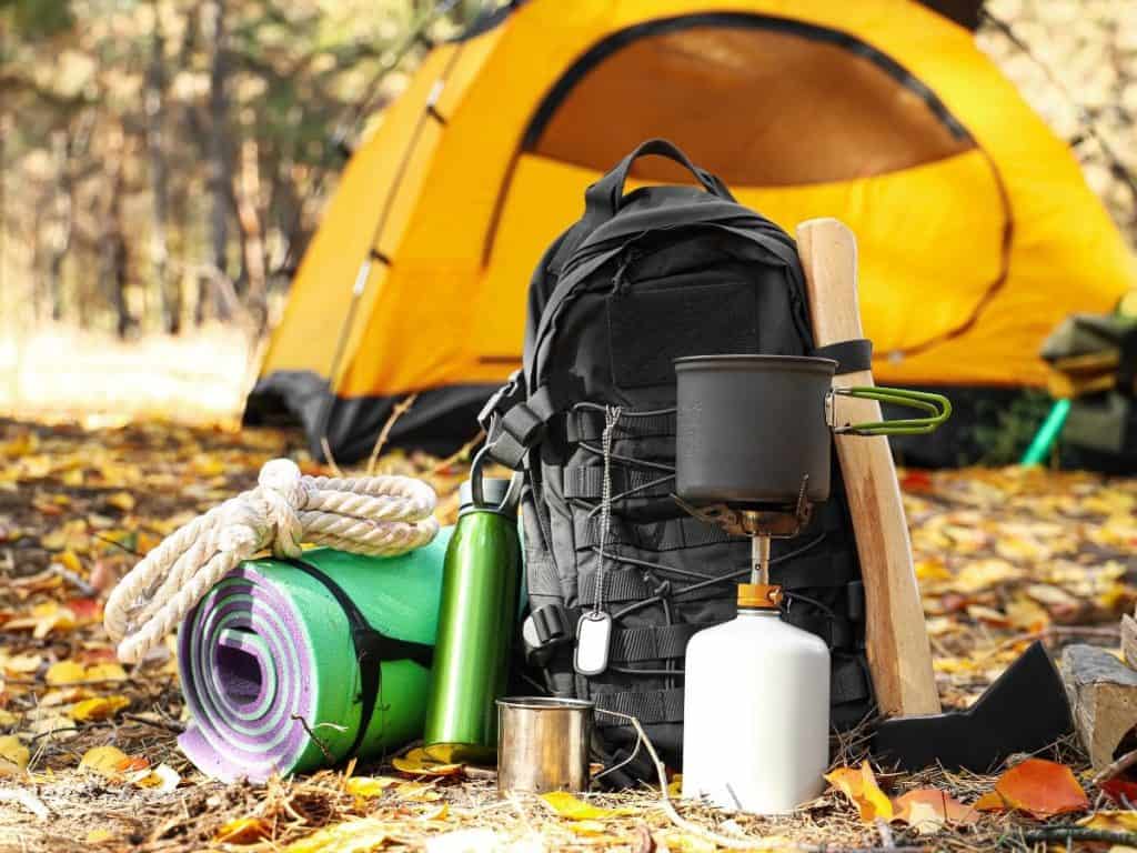 Backpack and camping gear set up beside a yellow tent in woodland, featuring a compact gas stove and pot, illustrating the best camping stove for outdoor cooking.