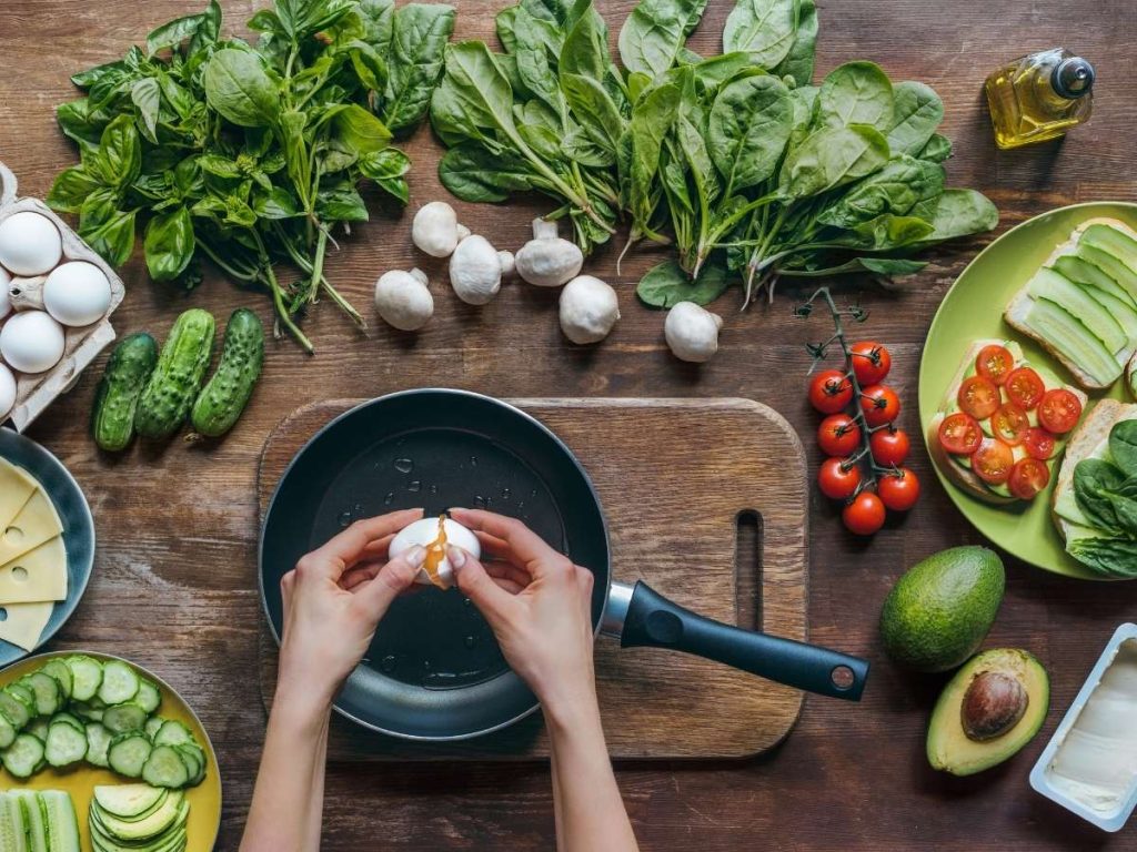 Overhead view of hands cracking an egg into a pan surrounded by fresh vegetables and simple ingredients on a wooden table, illustrating easy camping meals that can be cooked with minimal equipment.