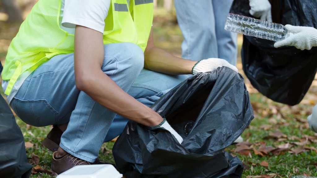 A volunteer wearing a high-visibility vest and gloves placing plastic waste into a rubbish bag during an outdoor clean-up - illustrating responsible waste management, packing out litter, and reducing environmental impact while camping.