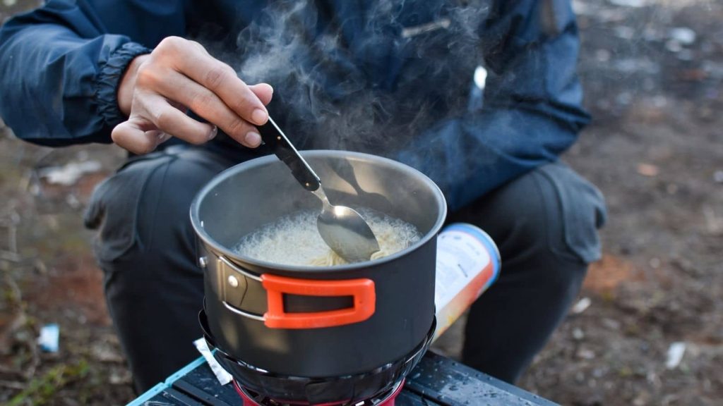 Close-up of a camper cooking on a portable gas stove outdoors, stirring food in a reusable metal pot - highlighting efficient, low-waste meal preparation and smarter food choices while camping to reduce environmental impact.