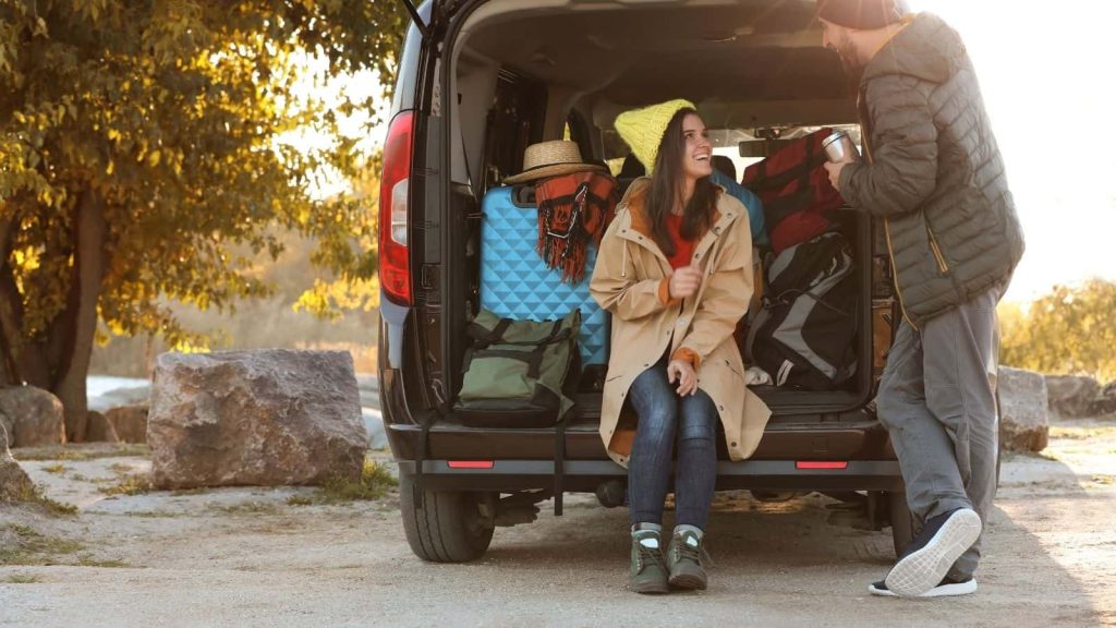 Couple preparing for a camping trip from the back of a van, surrounded by packed gear in warm evening light - representing travel choices, vehicle use, and how transportation impacts the overall environmental footprint of a camping trip.