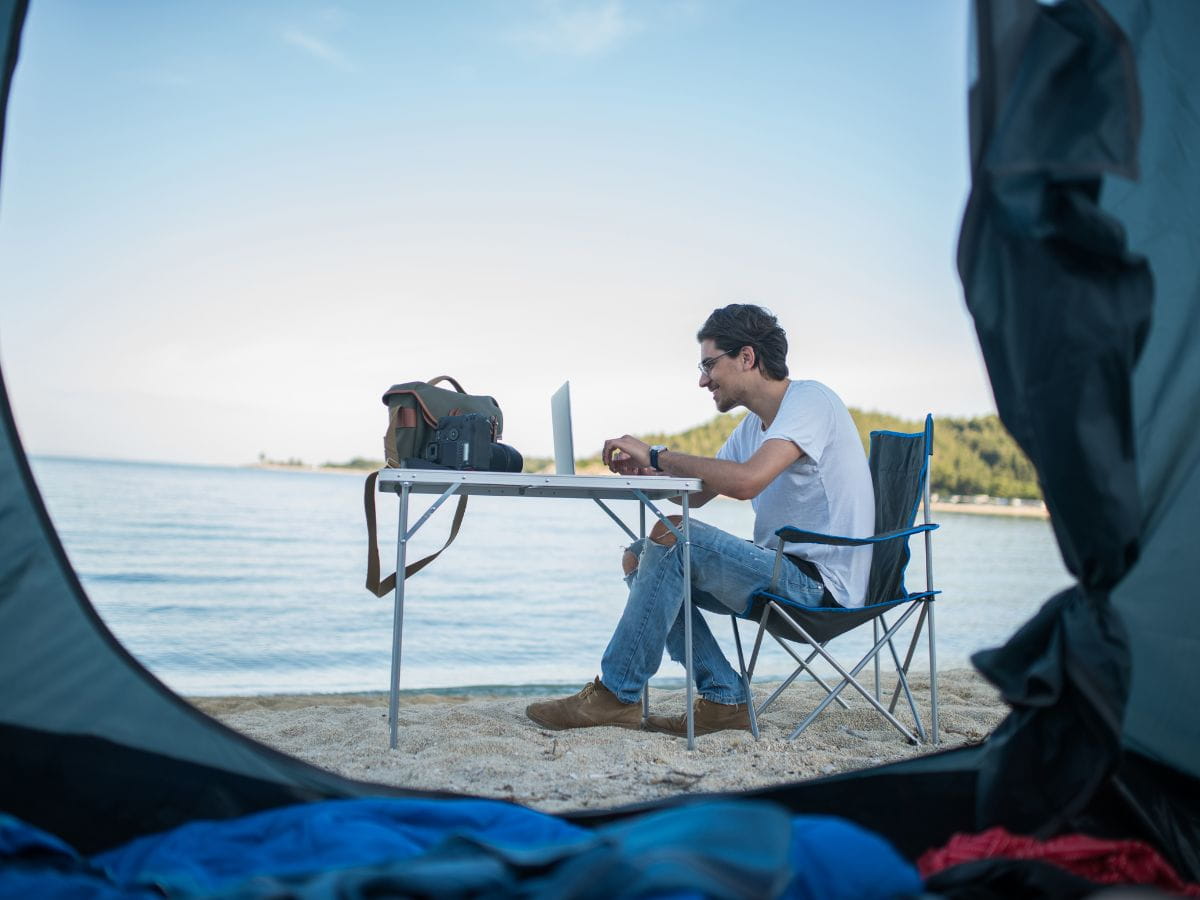 Person working on a laptop at a beach campsite, showing the reality of working remotely while camping outdoors.