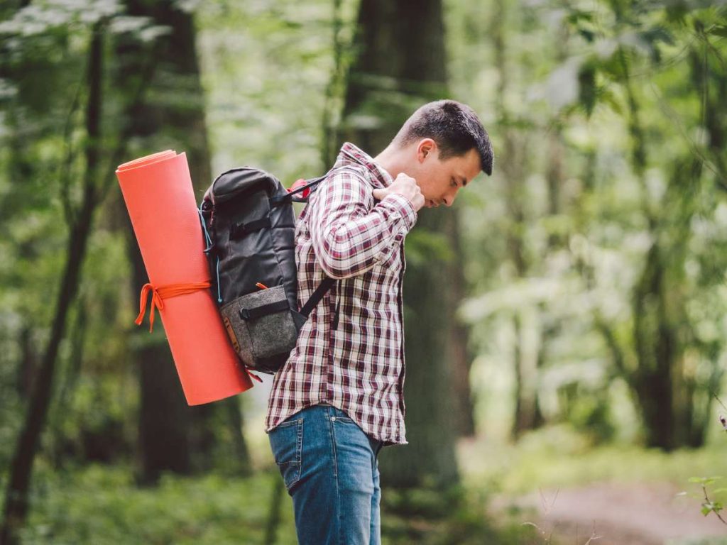 Person carrying camping gear backpack and sleeping mat illustrating carry-on vs checked luggage when flying with camping gear.
