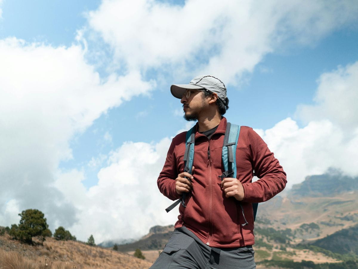 Male hiker wearing a lightweight fleece, cap and backpack beneath a cloudy mountain sky, illustrating the practical clothing layers to consider when deciding what to pack for a day hike.