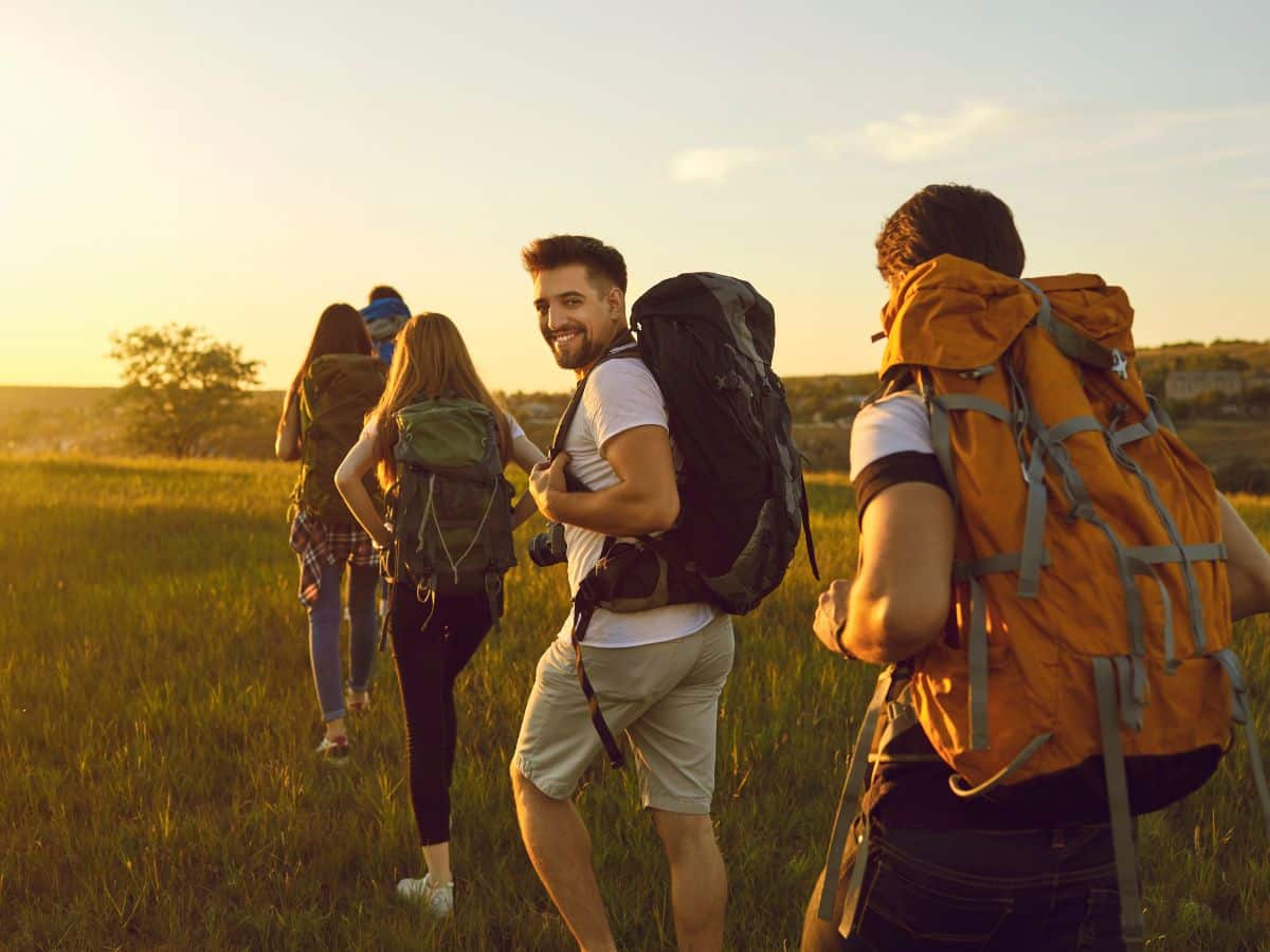 Group of hikers carrying large backpacks across open countryside in warm evening sunlight, illustrating sun protection and other easily forgotten items when deciding what to pack for a day hike.