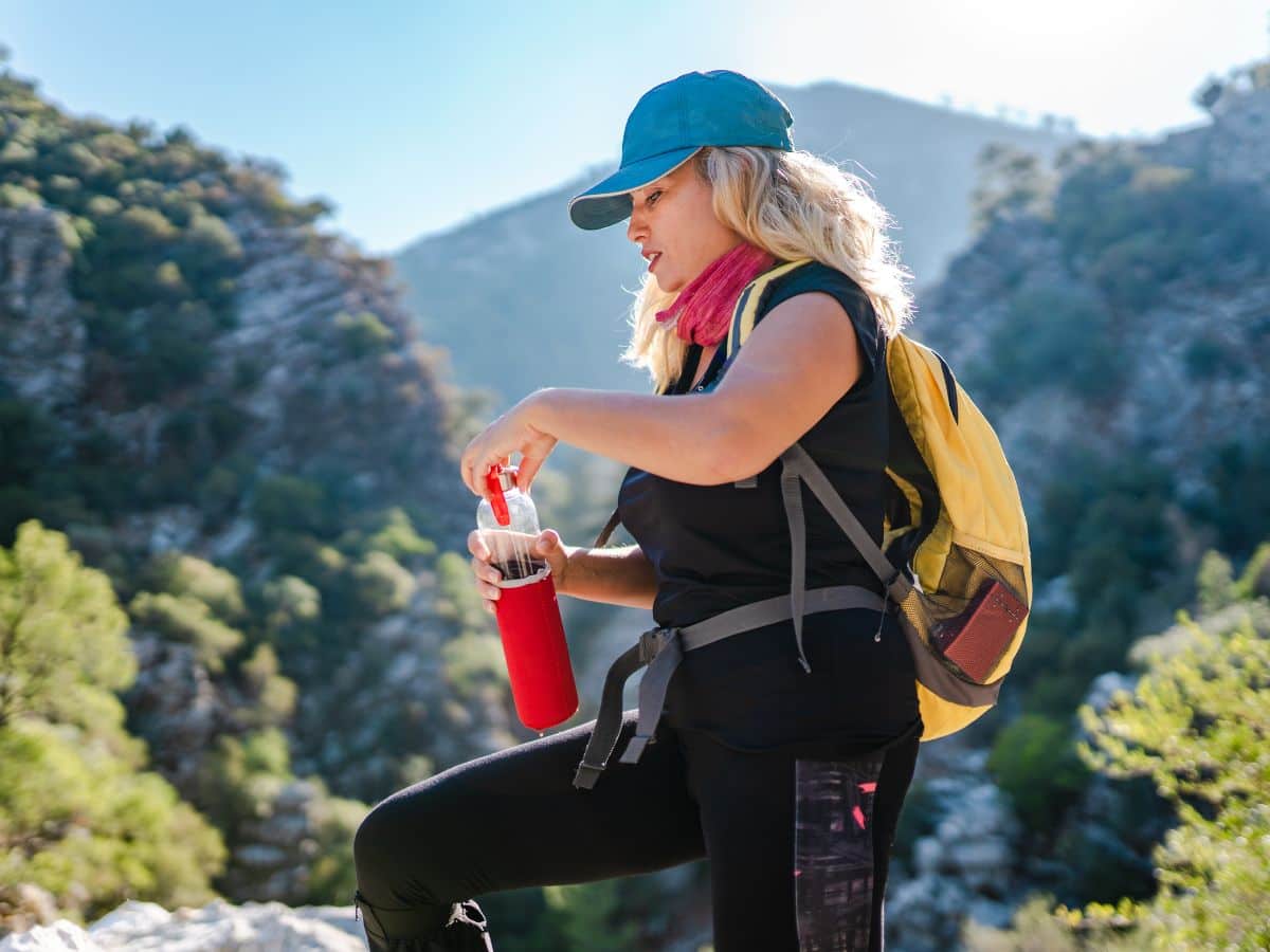 Woman hiker carrying a yellow daypack and opening a reusable water bottle on a sunny mountain trail, illustrating why water and food are essential when deciding what to pack for a day hike.