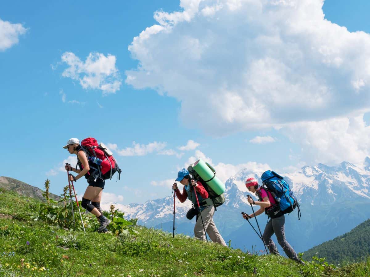 Three hikers carrying large backpacks, sleeping mats and trekking poles up a mountain slope, illustrating bulky equipment you can usually leave at home when deciding what to pack for a day hike.