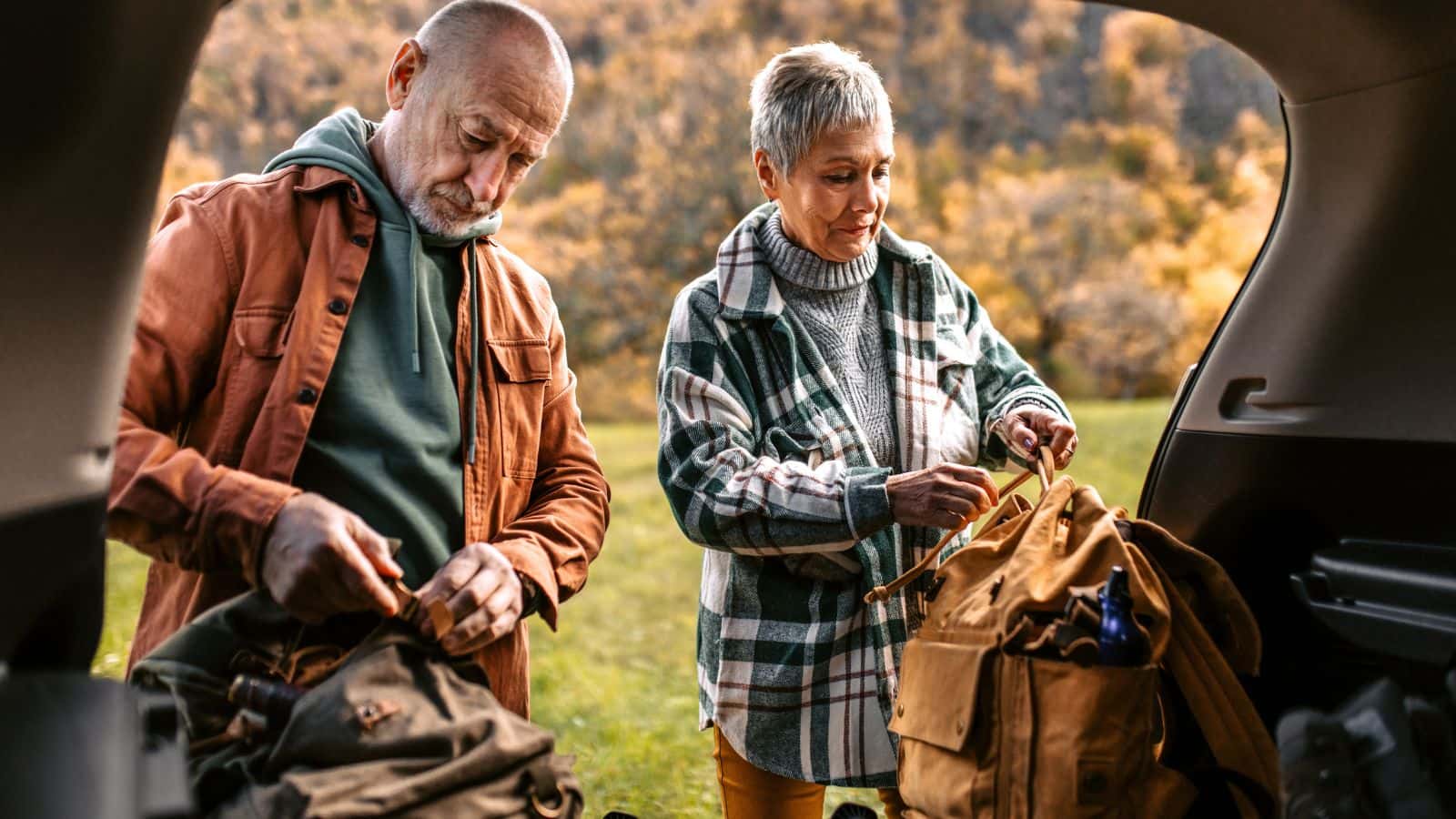 Older couple packing hiking bags beside an open car boot, illustrating the importance of careful pre-hike preparation when hiking with lower back pain.