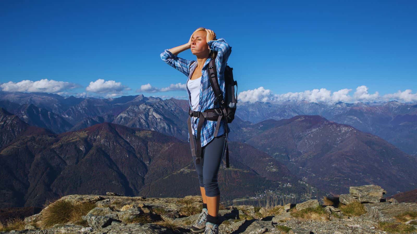 Woman hiker pausing on a mountain summit with her hands on her head, illustrating on-trail techniques for managing fatigue, posture and discomfort while hiking with lower back pain.