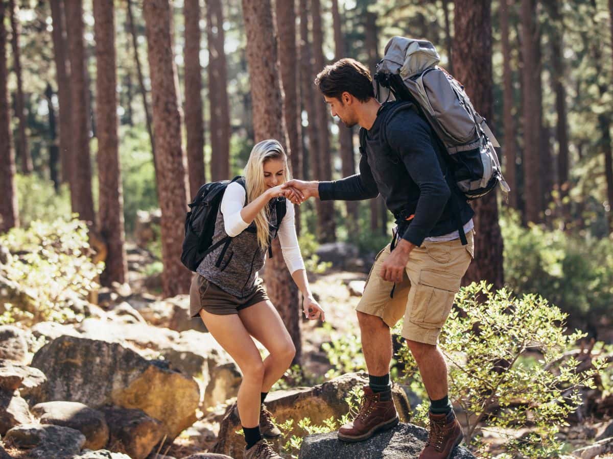 Two hikers carrying backpacks hold hands while crossing rocks on a forest trail, illustrating how arm position and restricted circulation can make hands swell up while hiking.