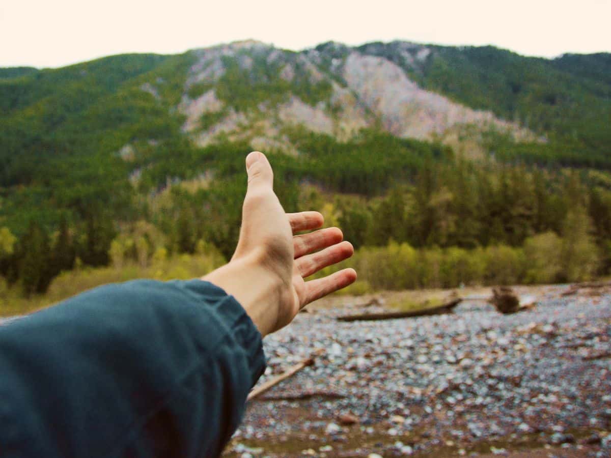 A hiker holds out an open hand towards a forested mountain, illustrating hidden trail factors that can make fingers and hands swell up while hiking.