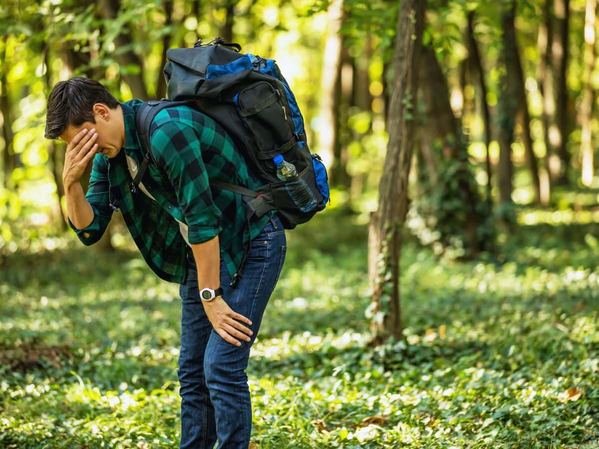Exhausted hiker bends forward with one hand covering his face on a woodland trail, illustrating when hands swell up while hiking alongside symptoms that may signal dehydration, heat illness or another medical concern.