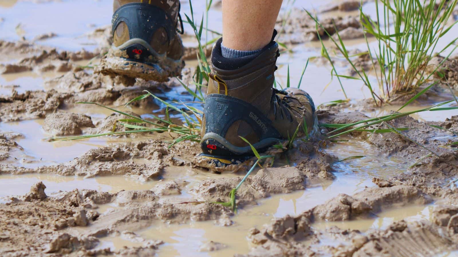 Close-up of hiking boots stepping through deep mud and puddles, showing careful foot placement and terrain reading on a wet, boggy trail.