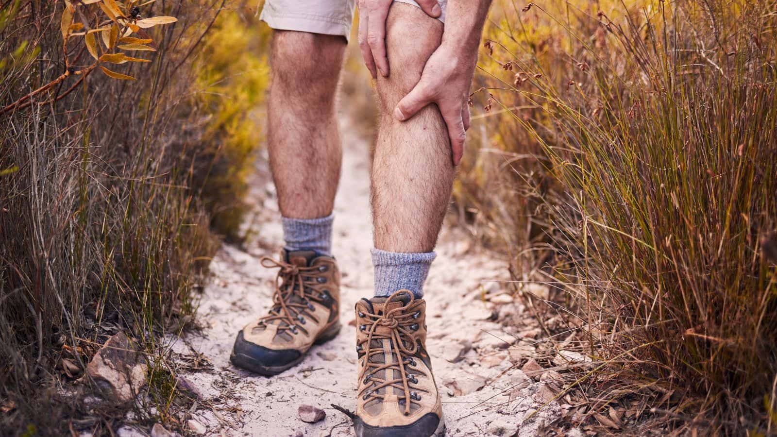 A close-up of a hiker clutching their leg just below the joint while standing on a dirt trail, illustrating the discomfort of knee pain when hiking downhill.