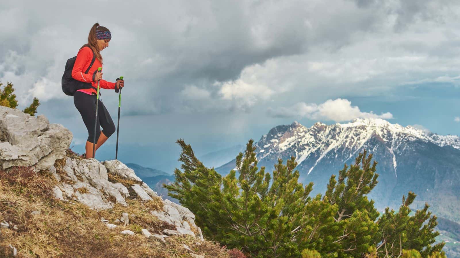 A female hiker using trekking poles to carefully step down a steep rocky mountain ledge, a technique to prevent knee pain when hiking downhill.